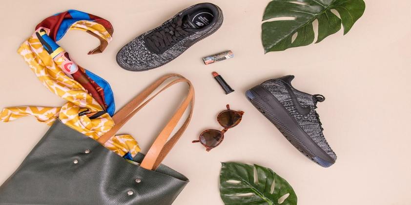 Unsplash. grey and brown bag with scarf, sunglasses, nike sneakers, and lip gloss on pink background with green leaves