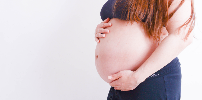 A pregnant woman stands against a white wall holding her belly